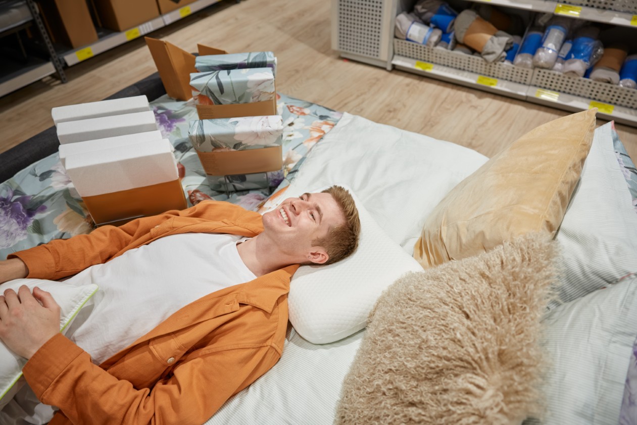 młody mężczyzna leżący na łóżku w sklepie, testujący ergonomiczną poduszkę ortopedyczną z pianki memory w otoczeniu ekspozycji materacy i pościeli